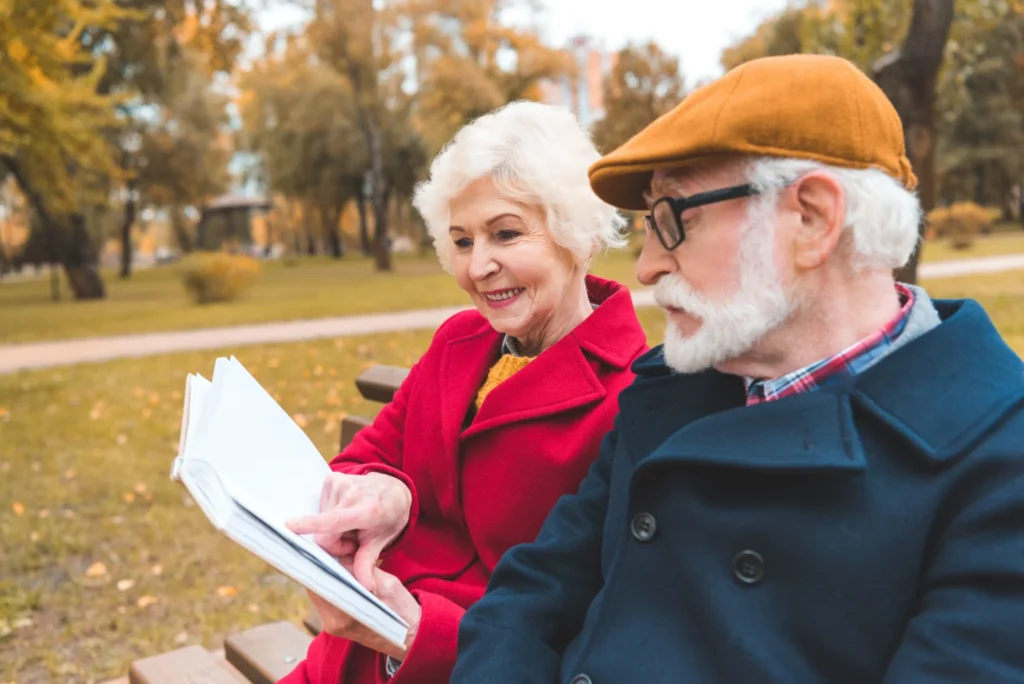 senior couple wearing coats sit outside on a park bench; woman points to a section of the book she's reading as man follows along.