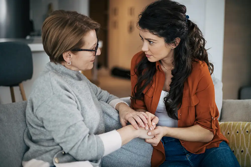 An adult daughter sits on a couch holding hands with her mother, one of the seniors at Clarendale Six Corners, providing emotional connection and caregiver support for elderly parents.